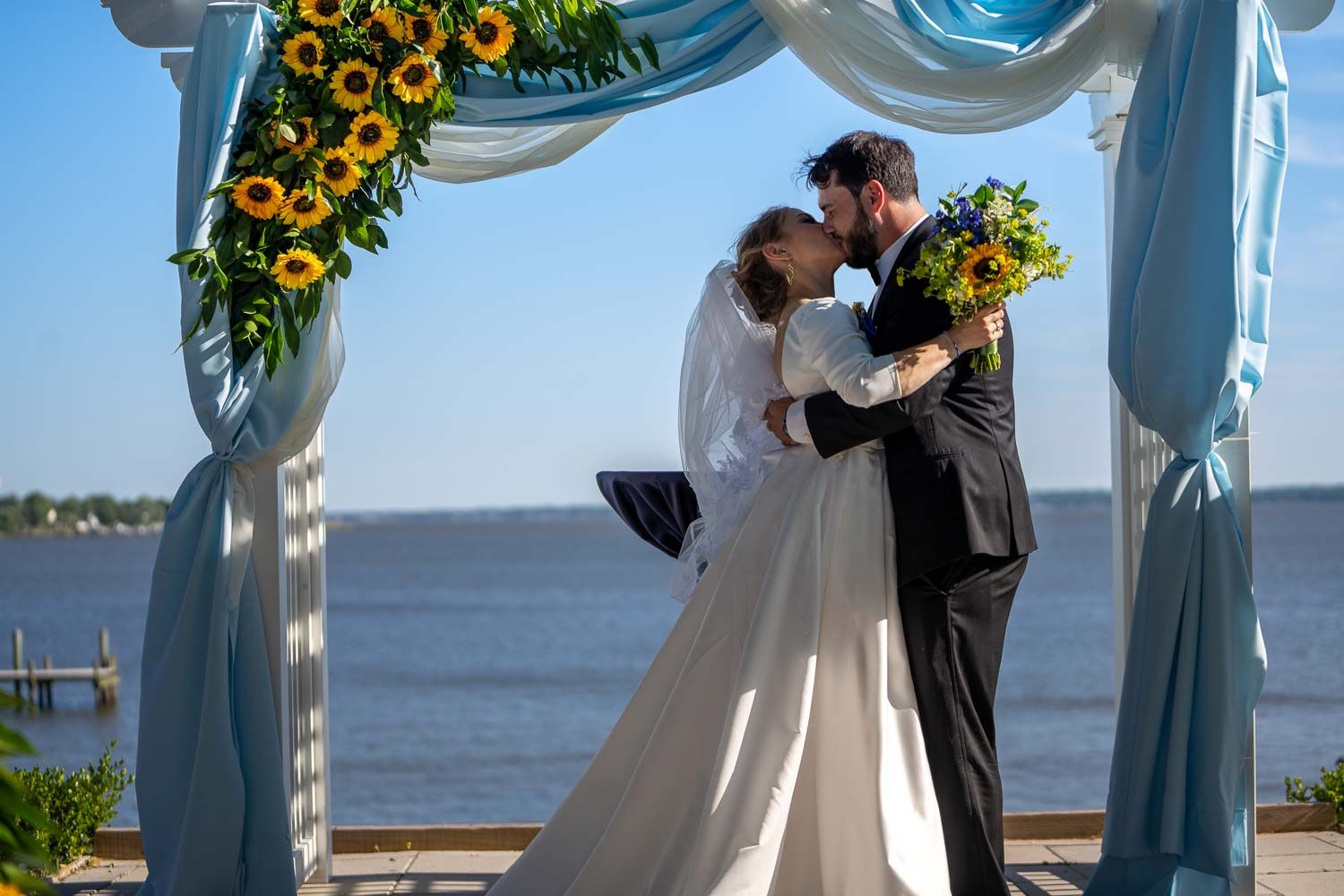 Bride and groom kissing under sunflower arch with blue draping and waterfront backdrop.