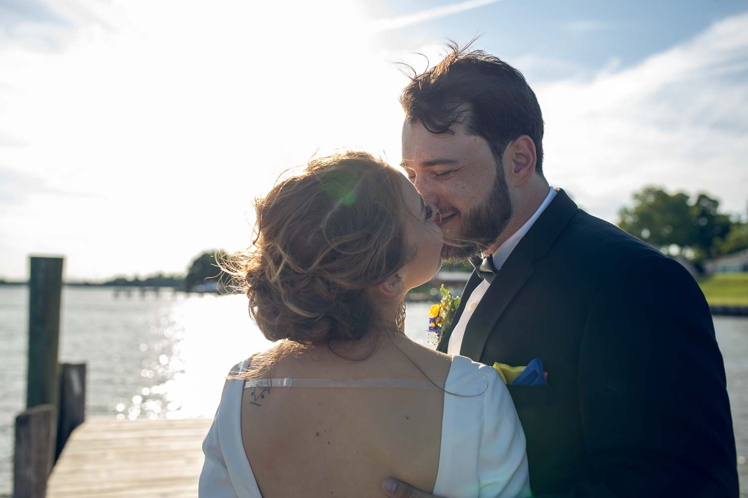 Newlyweds share a tender kiss on a sunlit dock with water and bokeh background.