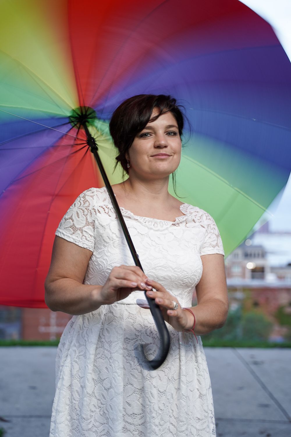 Woman in white lace dress holding a colorful rainbow umbrella outdoors with a soft smile on her face.