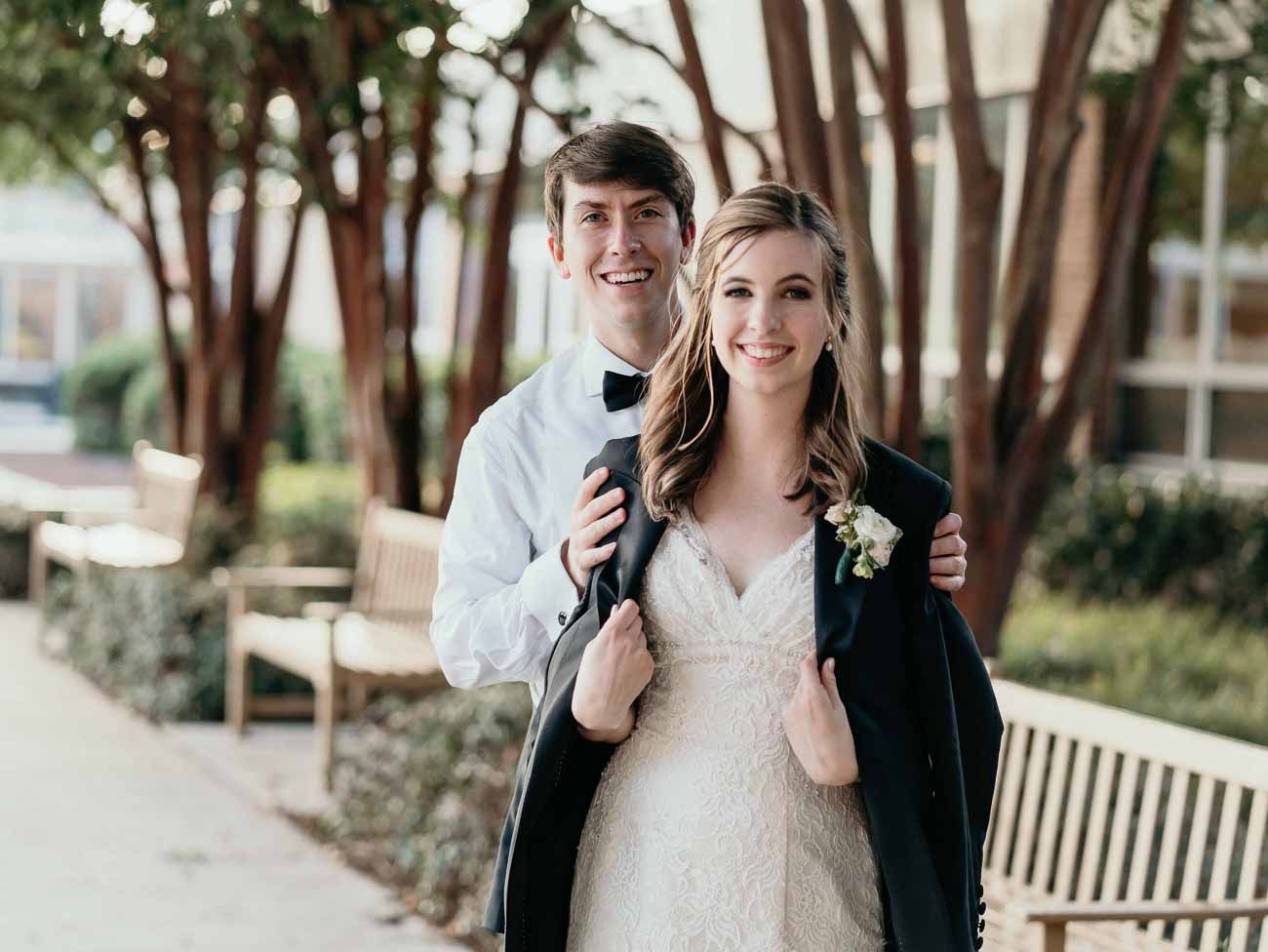 Groom wraps his tuxedo jacket around bride's shoulders as they pose outdoors near wooden benches.