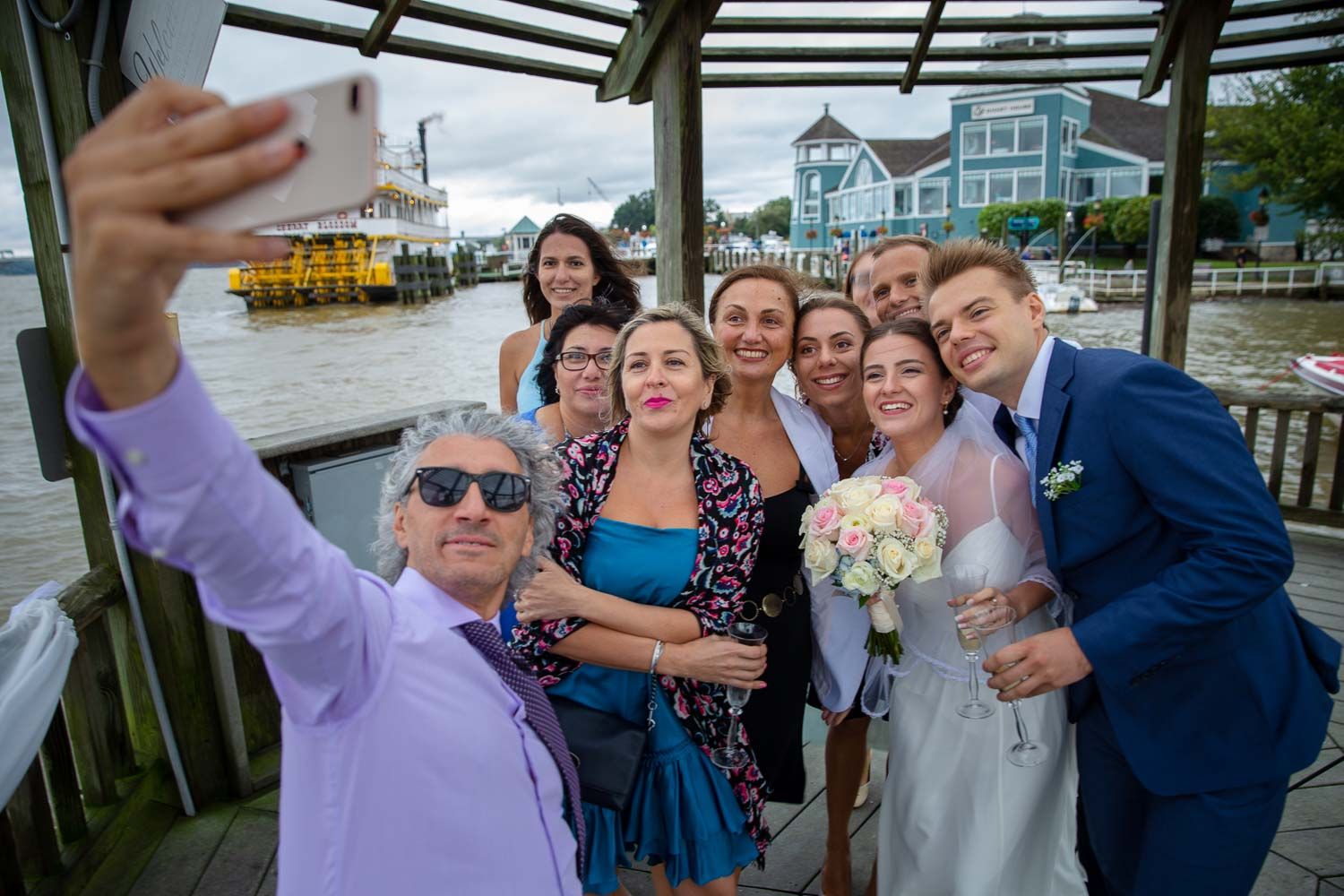 Wedding group selfie with smiling guests, bride with bouquet, and groom near scenic waterfront.