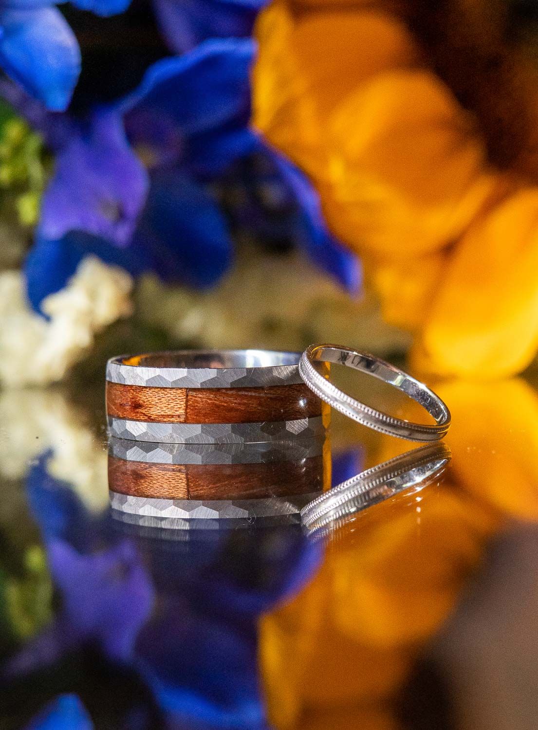Two wedding bands, one wood and hammered metal, one slim silver, reflected on a glossy surface with blue and yellow flowers.