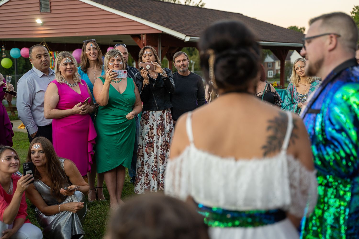 Wedding guests smile and photograph bride and groom during outdoor backyard wedding ceremony at sunset.