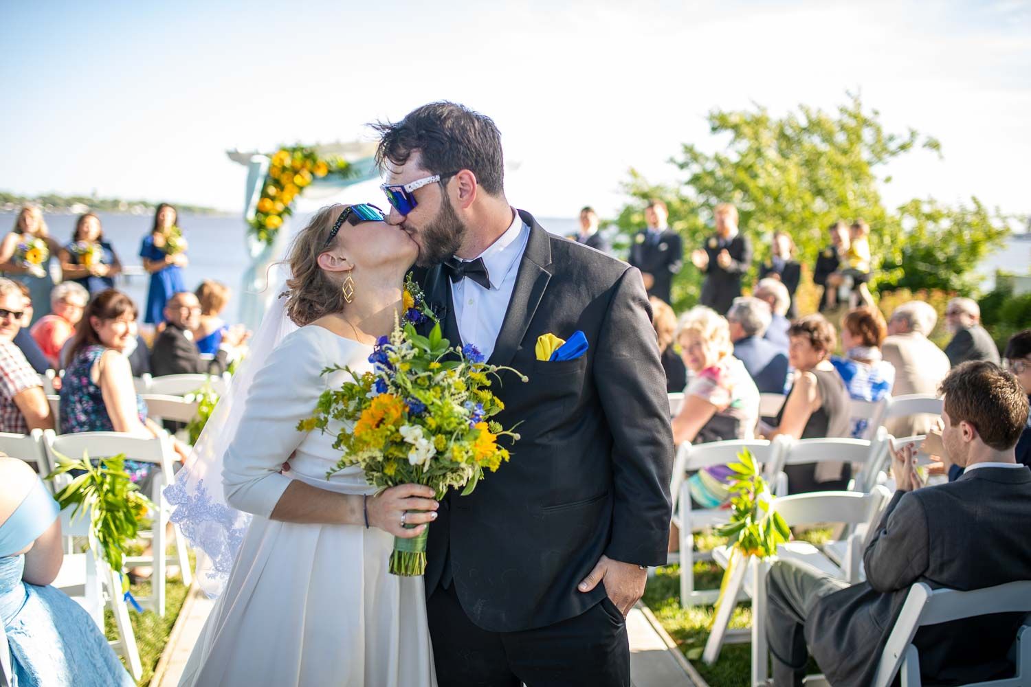 Bride and groom sharing first kiss wearing sunglasses at outdoor wedding ceremony with guests.