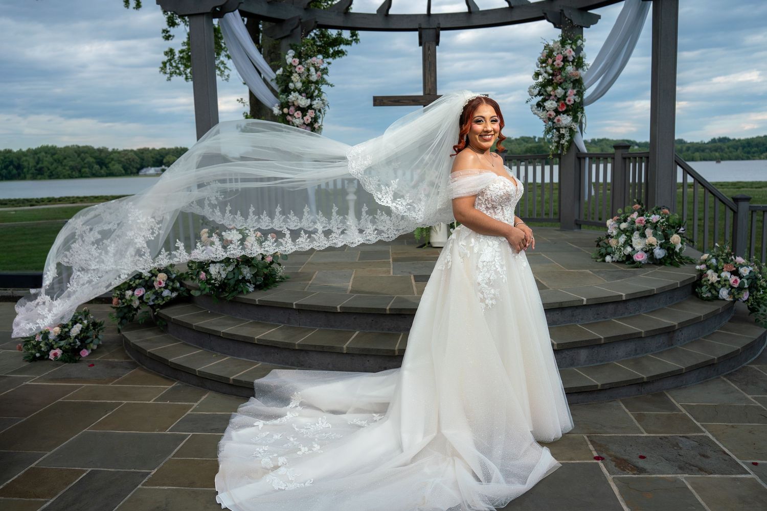 Bride in off-shoulder white gown with flowing veil at outdoor waterfront wedding venue with floral arch.