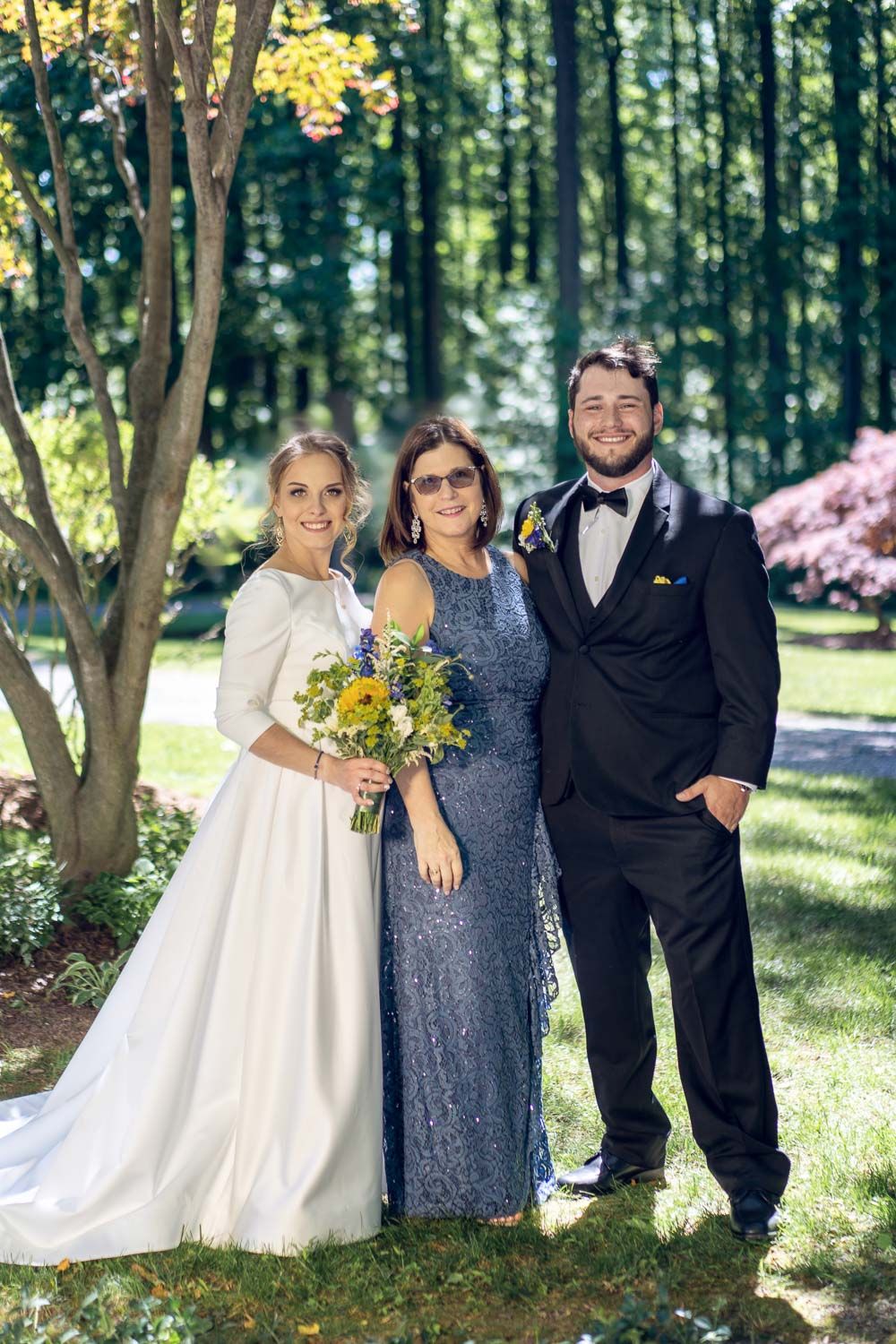 Bride and groom posing with woman in blue sequin dress holding sunflower bouquet outdoors.