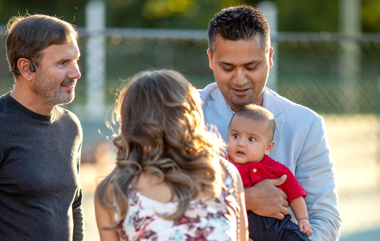 Man holding baby in red outfit interacting with couple at a warm outdoor social gathering.
