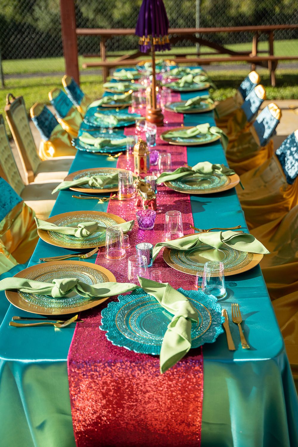 Luxurious Moroccan-themed banquet table with teal linen, pink sequin runner, gold chargers, and green napkins outdoors.