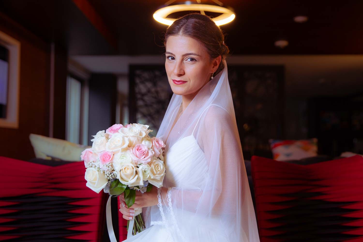 Elegant bride in white wedding dress and veil holding a pink and white rose bouquet indoors.
