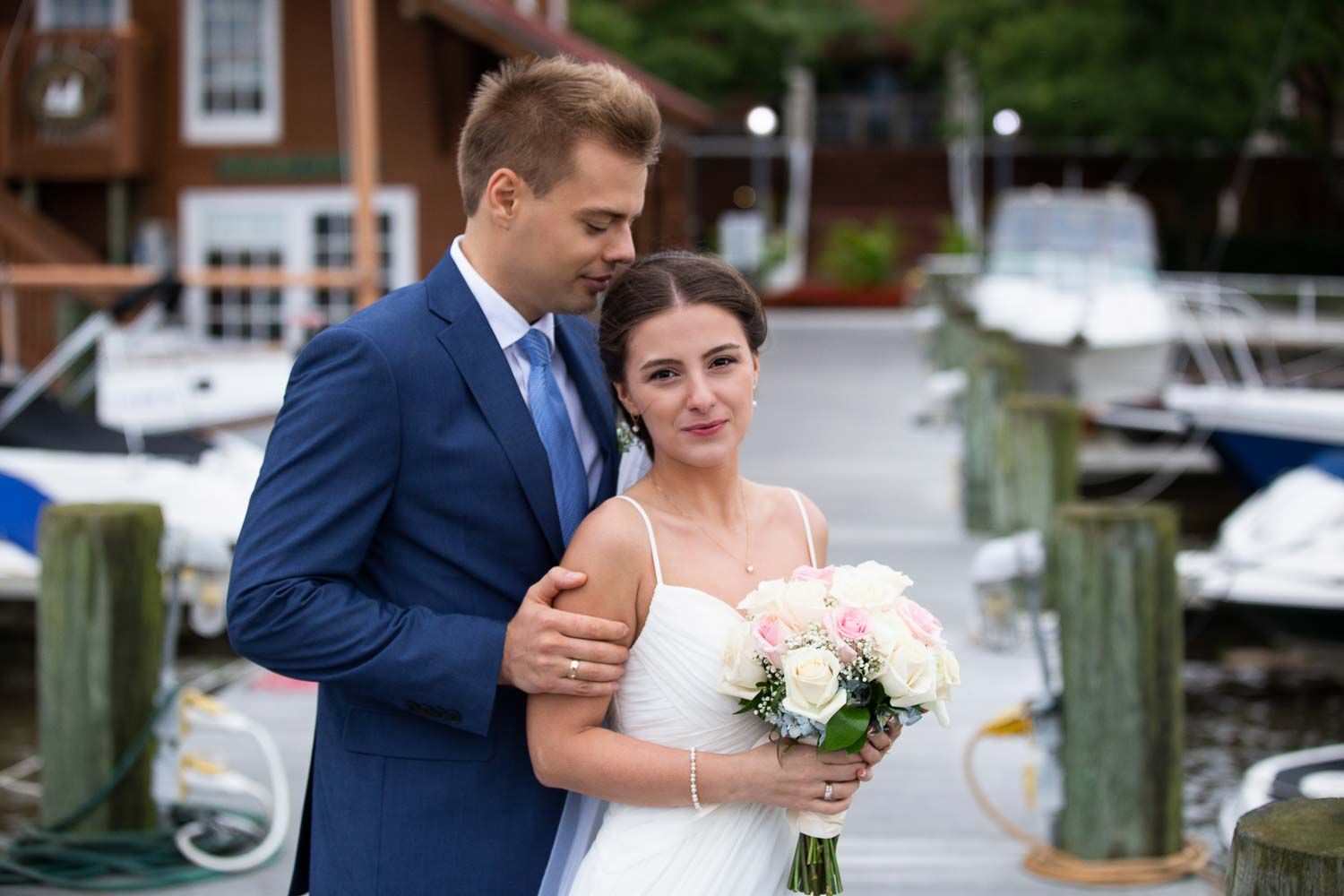 Bride and groom posing at marina with pink bouquet, boats and dock visible in background.