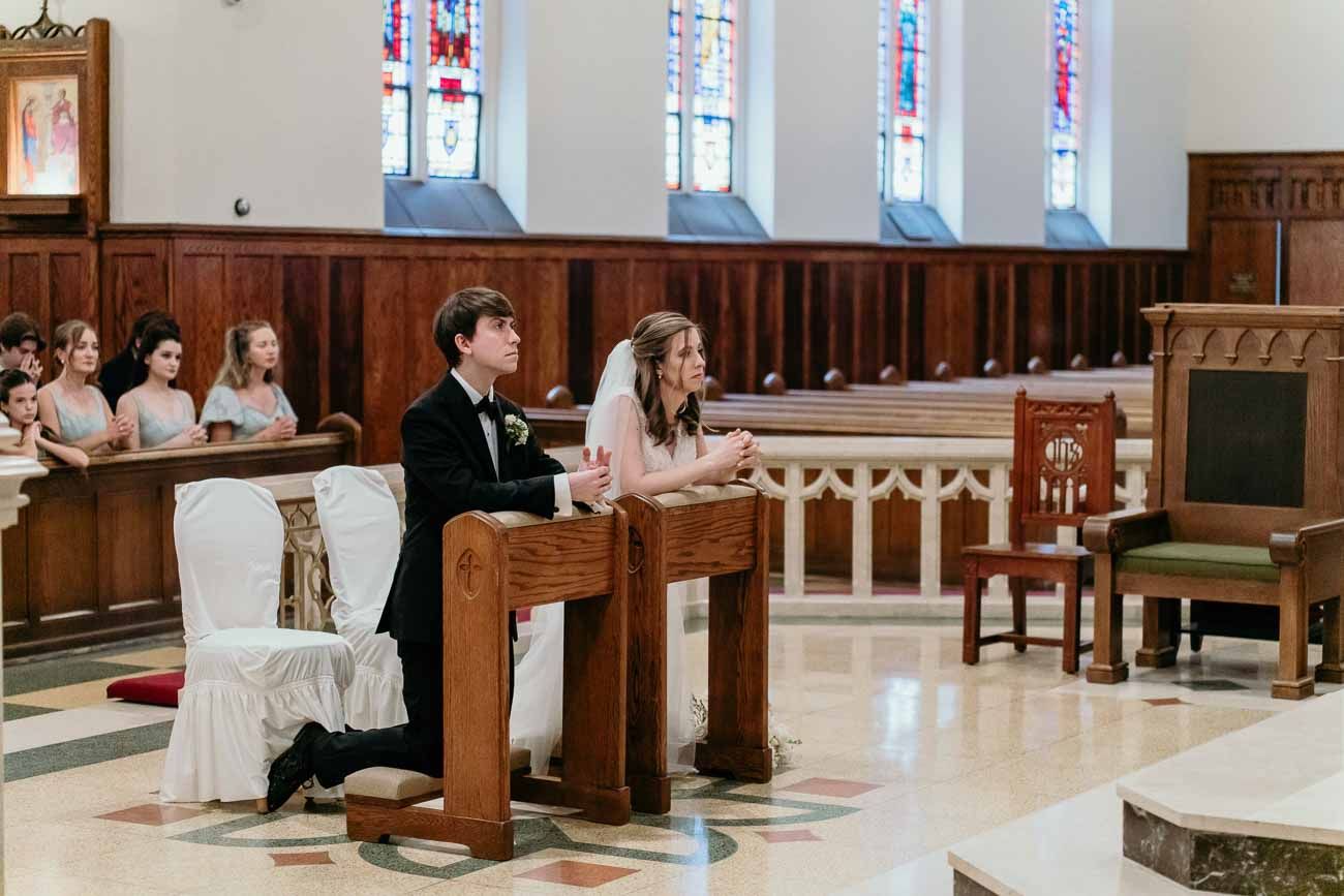 Bride and groom kneel at the altar during their Catholic wedding ceremony with guests seated in pews.