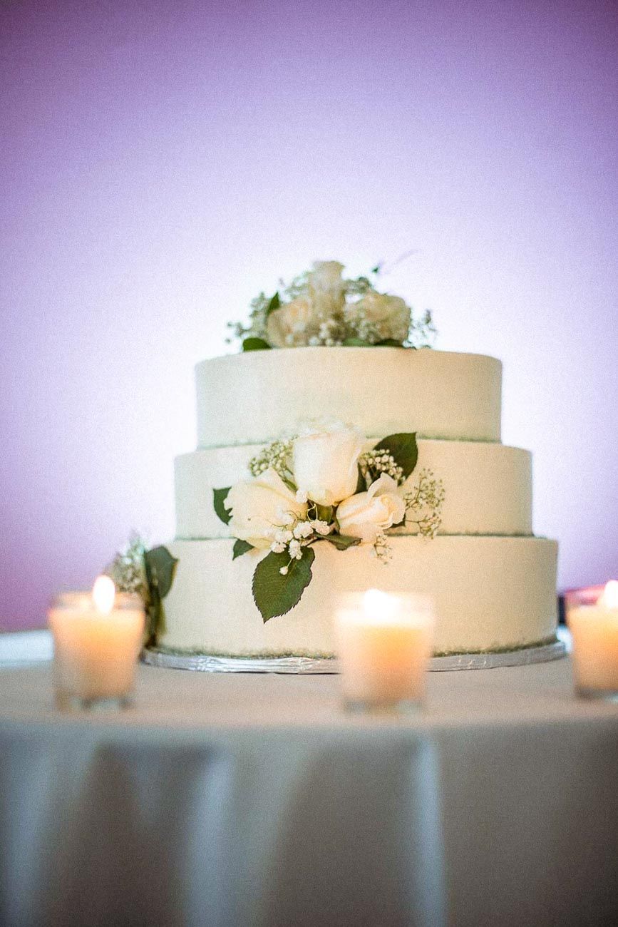 Elegant three-tier white wedding cake adorned with white roses and greenery, surrounded by glowing candles.