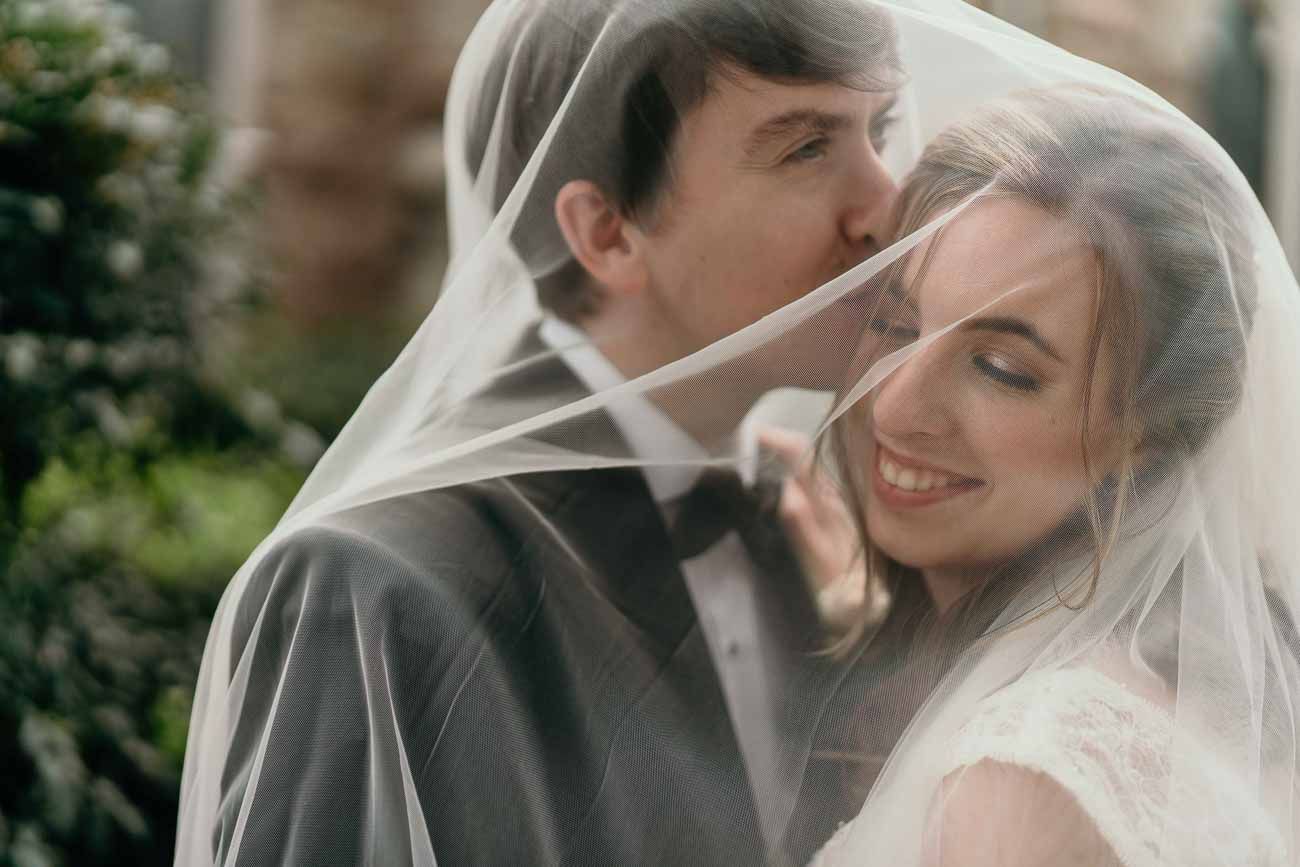 Groom nuzzles smiling bride under her flowing white veil outdoors on their wedding day.