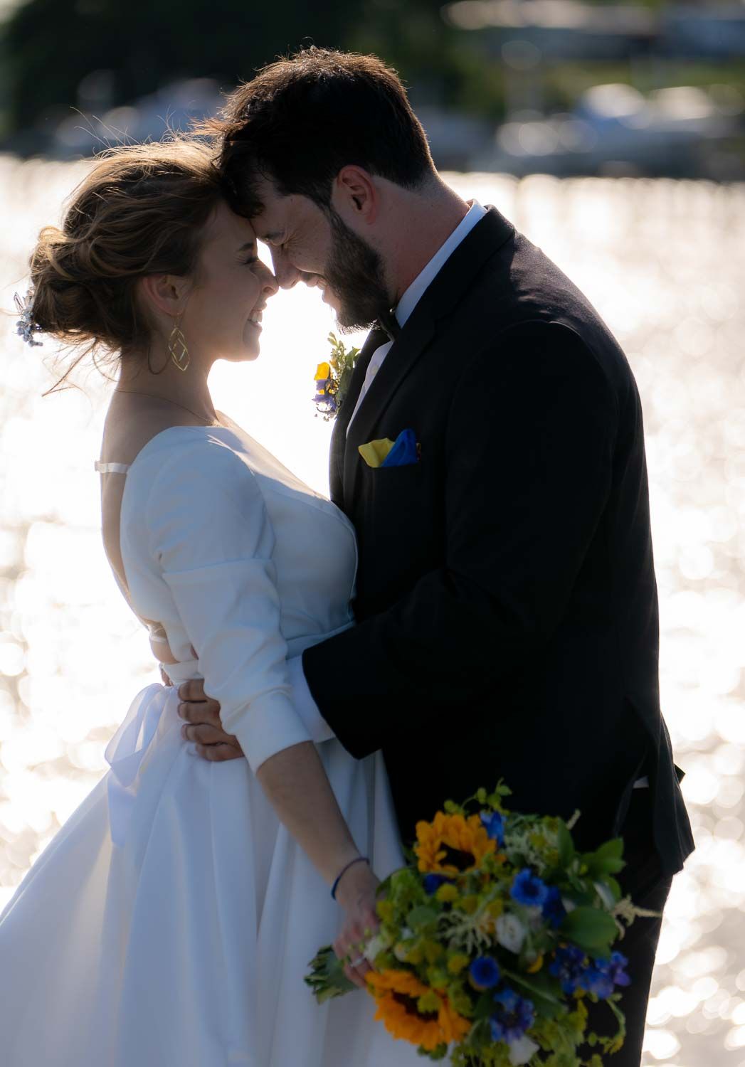 Bride and groom touching foreheads romantically by sparkling waterfront, bride holding sunflower bouquet.