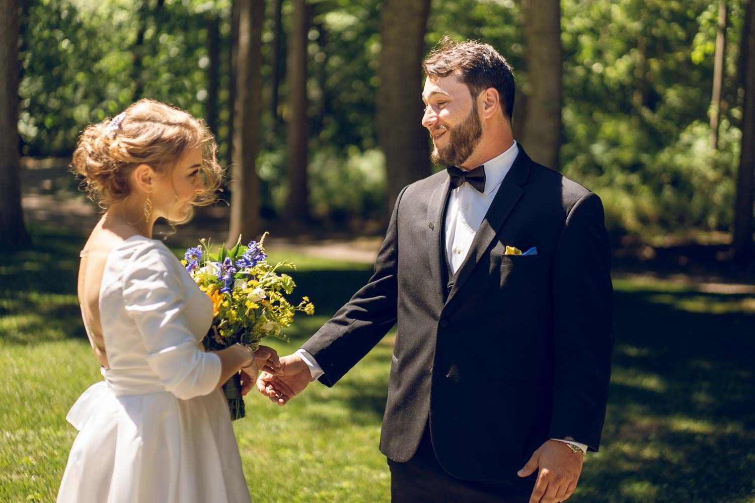 Bride and groom holding hands exchanging smiles while holding wildflower bouquet in sunny garden.