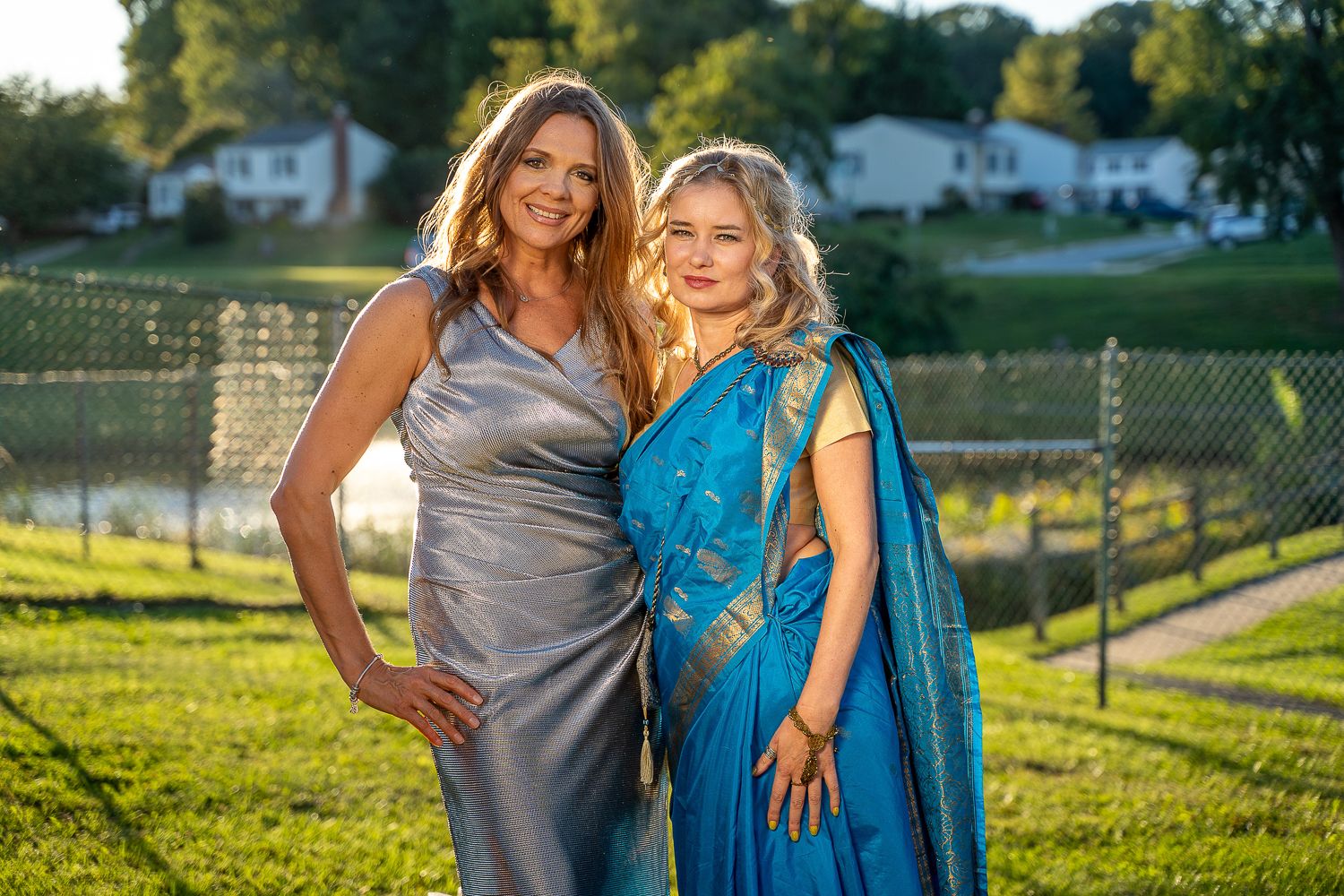 Two women in formal attire, one in silver gown and one in blue sari, posing outdoors at sunset.