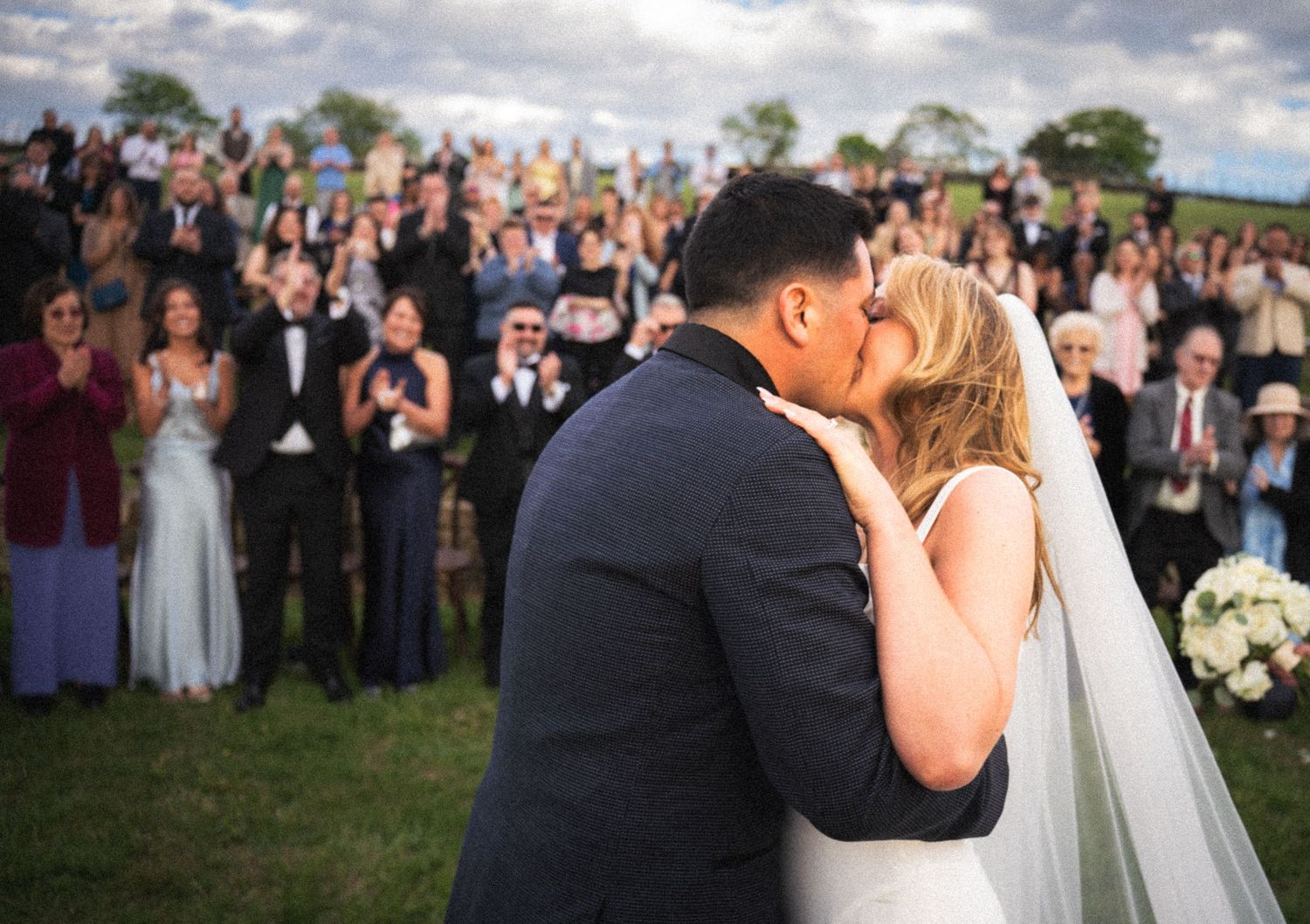 Bride and groom share first kiss at outdoor wedding ceremony with cheering guests applauding in background.