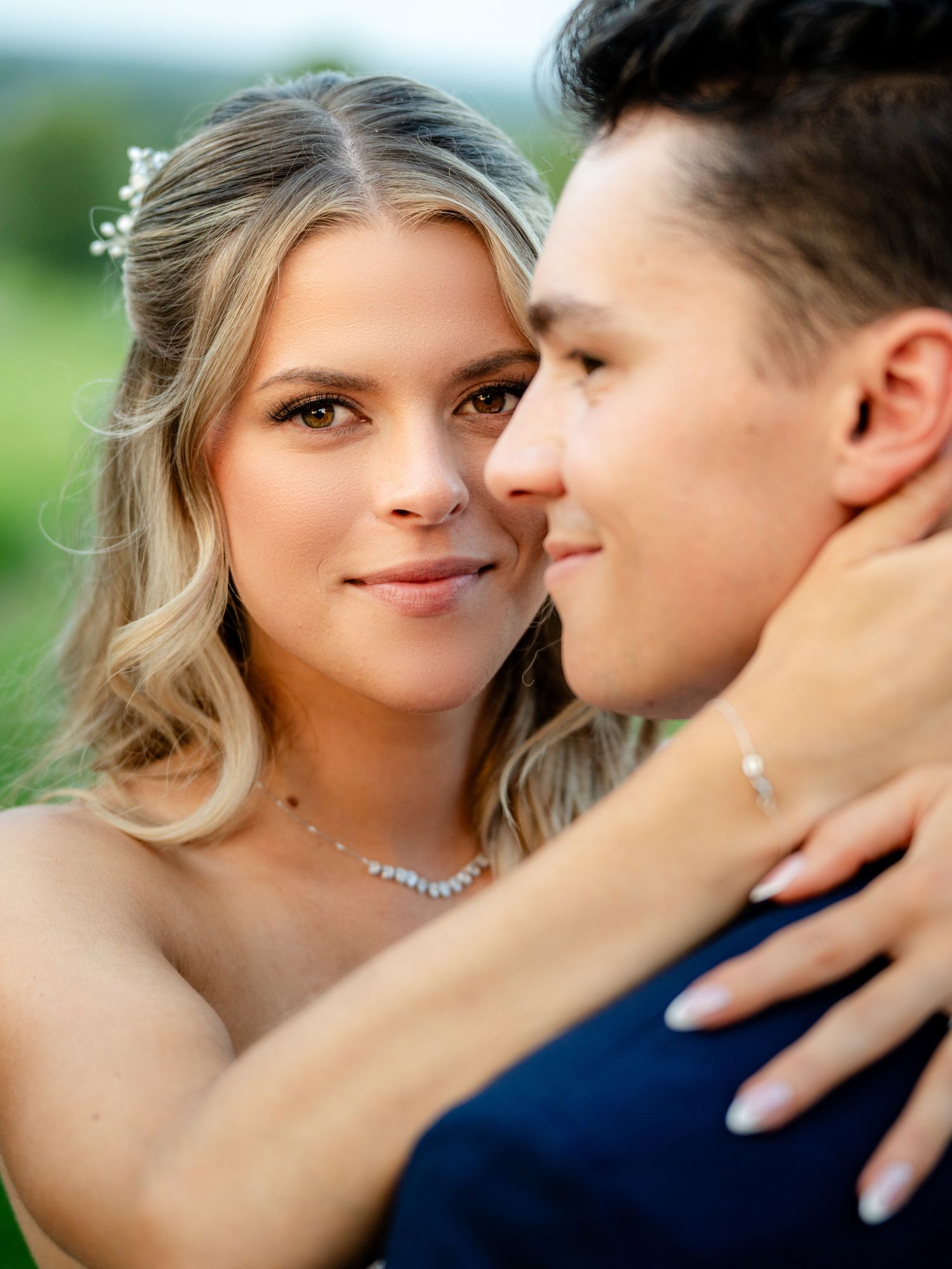 Bride with blonde hair and diamond necklace embraces groom in navy suit outdoors at romantic wedding portrait.