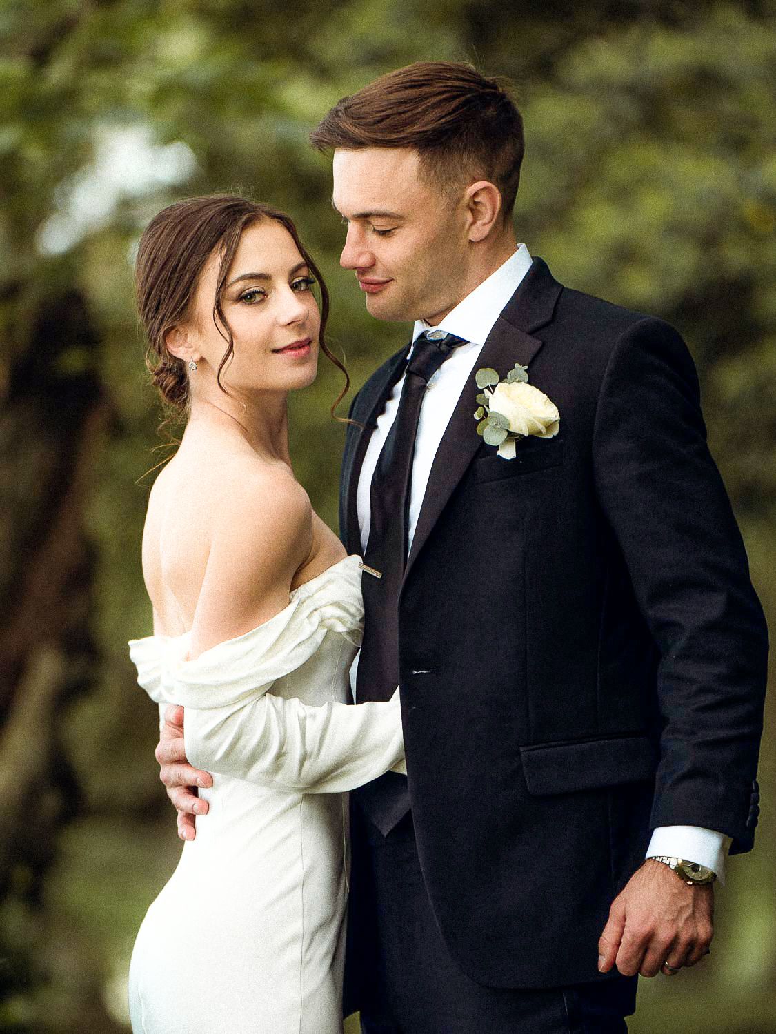 Elegant bride in off-shoulder gown poses with groom in black suit outdoors.