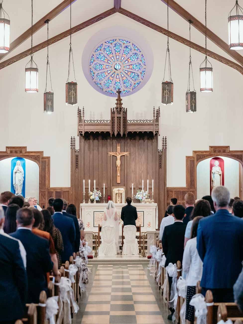 Wedding couple stands at altar of beautiful Catholic church with rose window and crucifix.