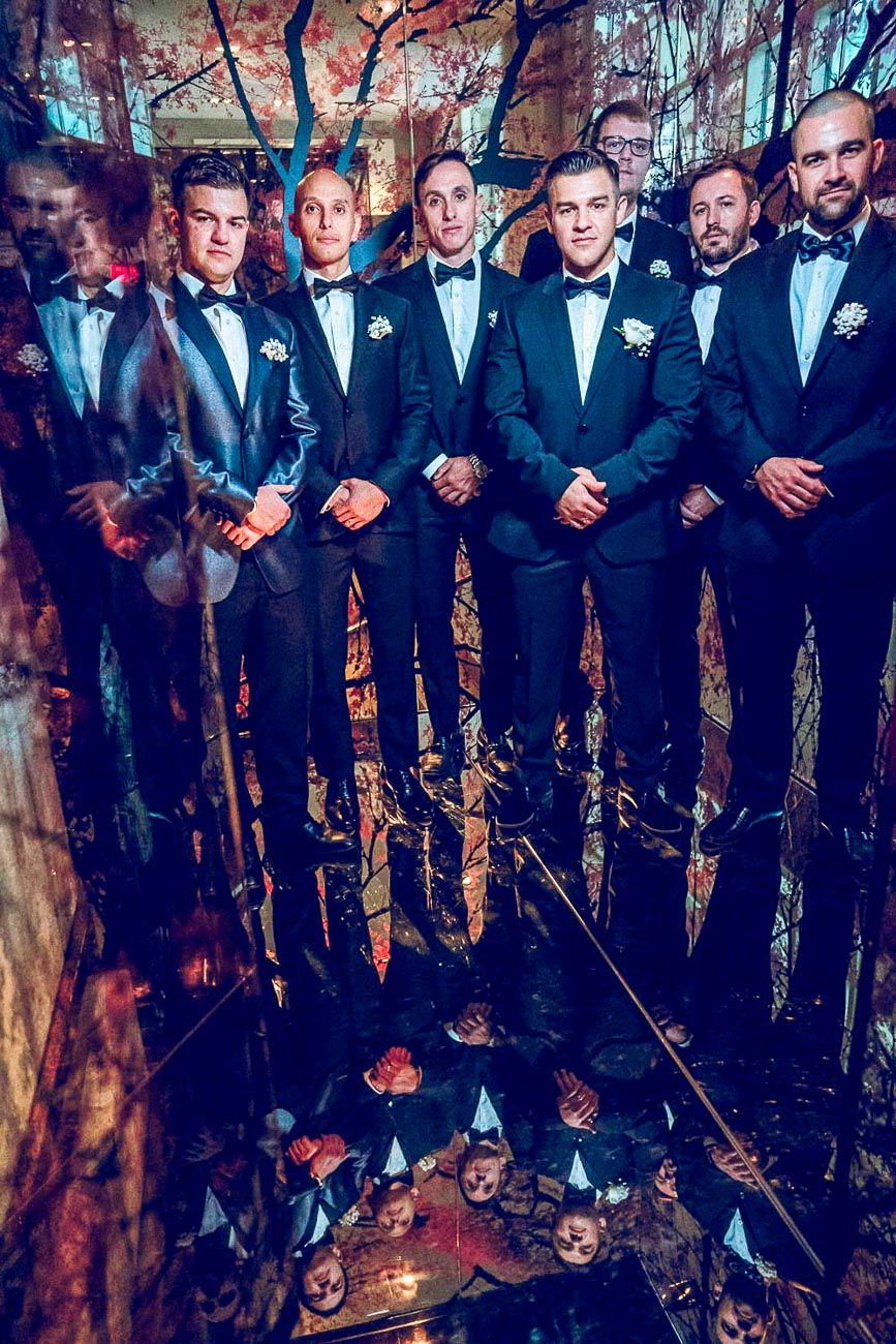 Seven groomsmen in black tuxedos with boutonnieres posing in stunning mirrored wedding venue.