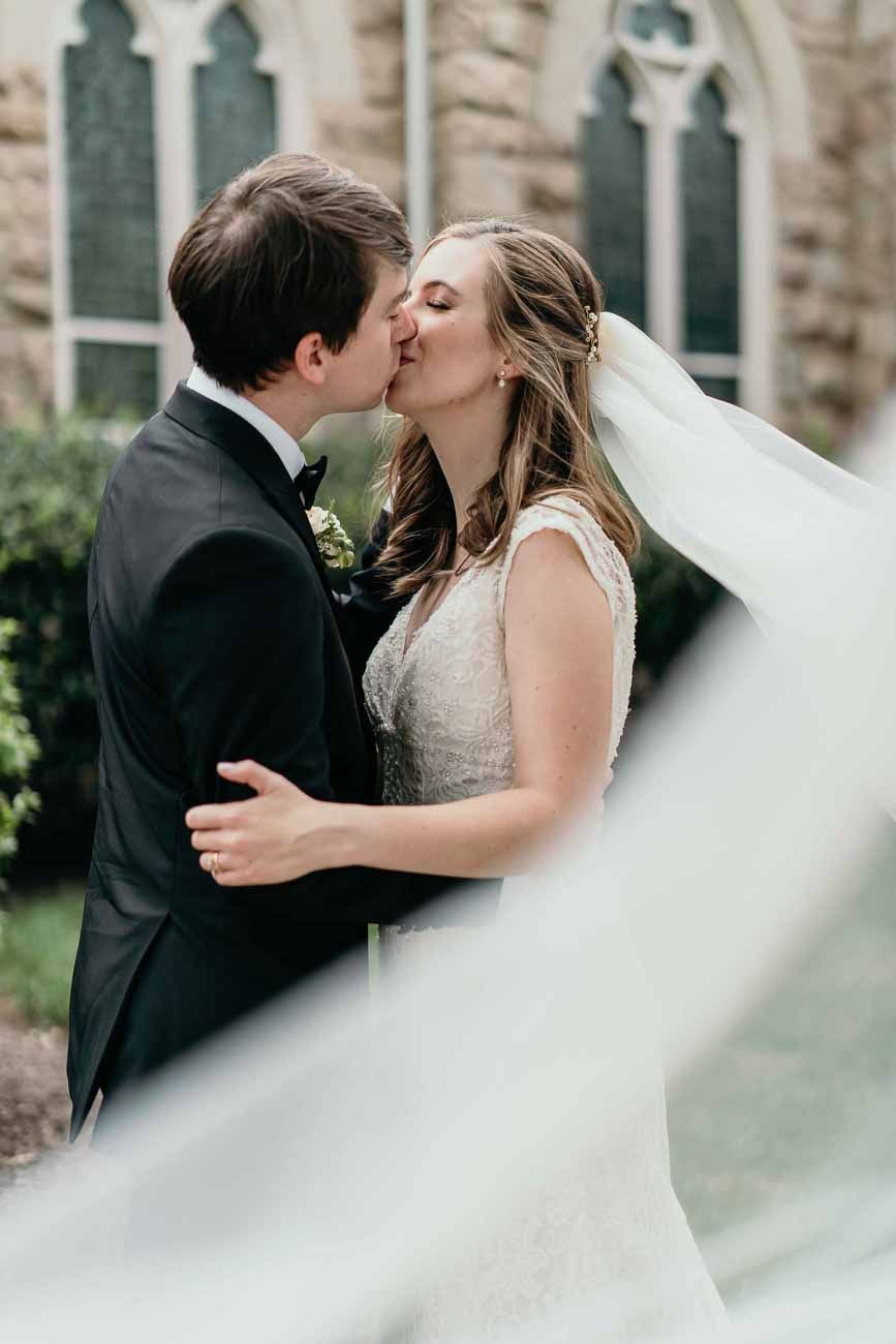 Bride and groom share a romantic kiss outdoors with veil flowing near a historic stone church.
