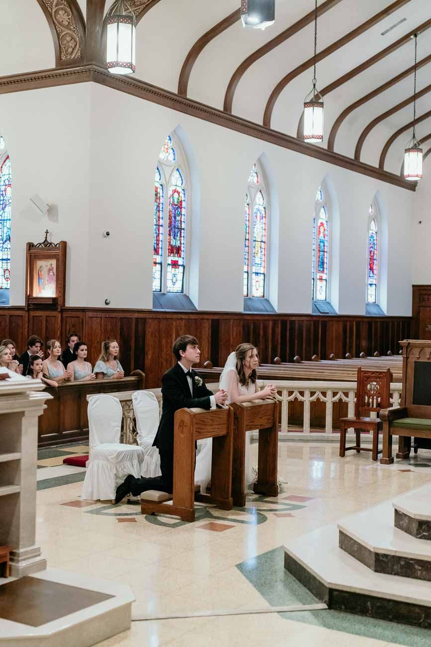 Bride and groom sit at kneeler before the altar in a beautiful Catholic church with stained glass windows.