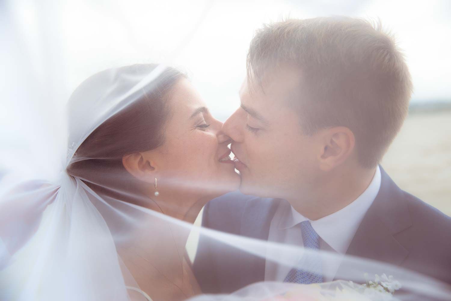 Romantic close-up of bride and groom sharing a kiss beneath a soft, flowing white veil.