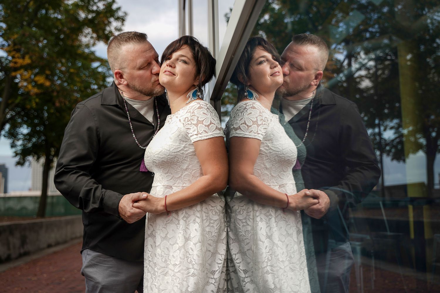 Groom kissing bride on cheek with glass reflection, bride in white lace dress wearing turquoise earrings outdoors.