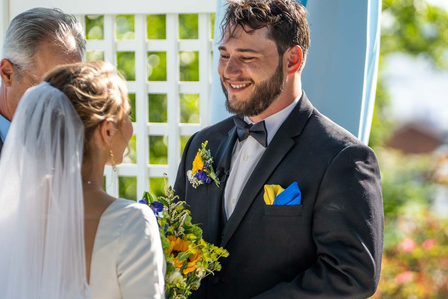 Groom in black tuxedo with sunflower boutonniere smiles joyfully at bride during outdoor ceremony.