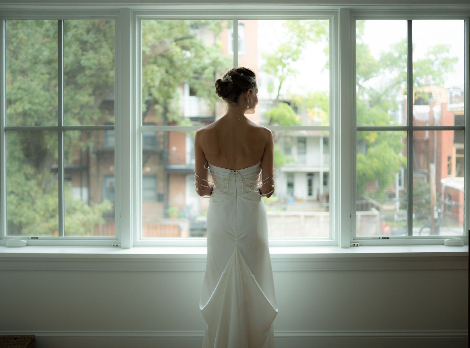 Bride in a strapless white wedding gown standing at a large window gazing outside before the ceremony.