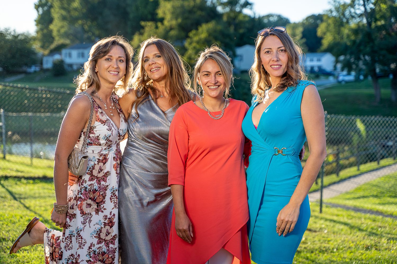 Four smiling women in elegant dresses posing together outdoors at a sunset celebration event.