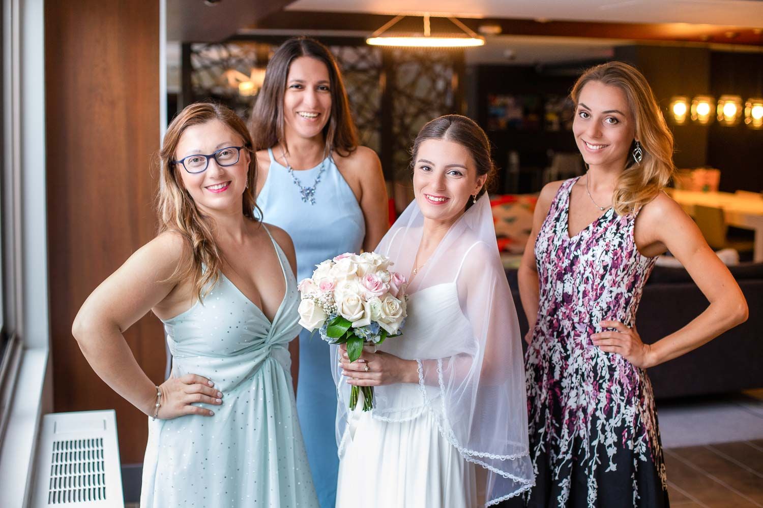 Bride in white gown and veil posing with three bridesmaids in elegant dresses holding a rose bouquet.
