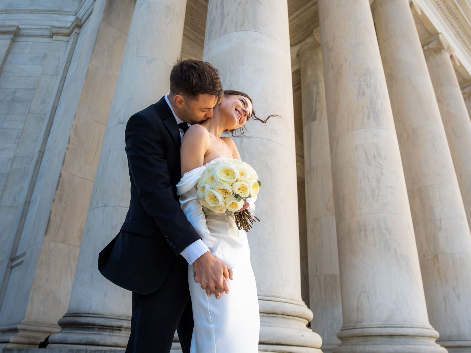Bride and groom embrace tenderly among grand marble columns with white floral bouquet.