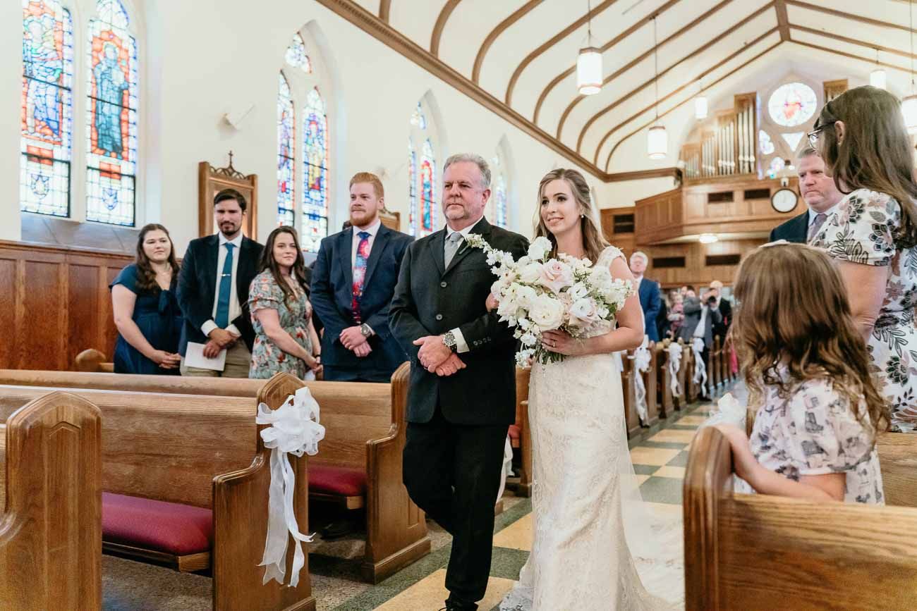 Bride carrying white bouquet walks down church aisle with father past smiling wedding guests.