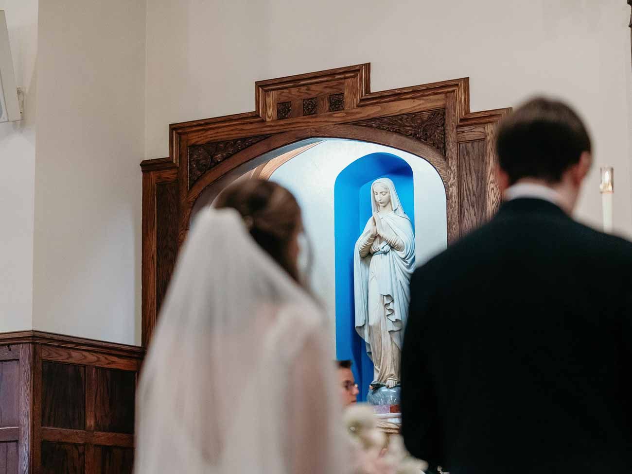Bride and groom stand before a white Virgin Mary statue in a blue niche during a Catholic wedding ceremony.