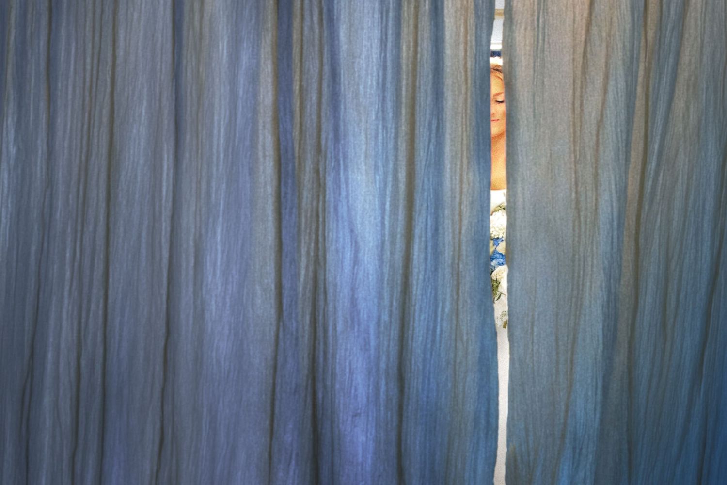 Close-up of flowing blue-gray curtains with a narrow gap revealing colorful objects behind the fabric.