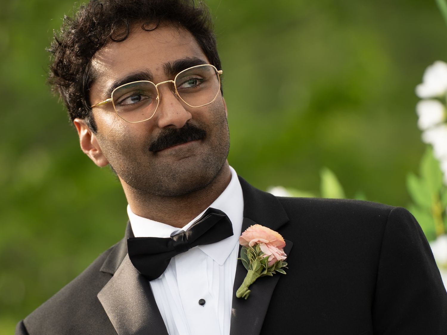 Young man in tuxedo with bow tie and boutonniere, wearing glasses, smiling outdoors at a formal event.
