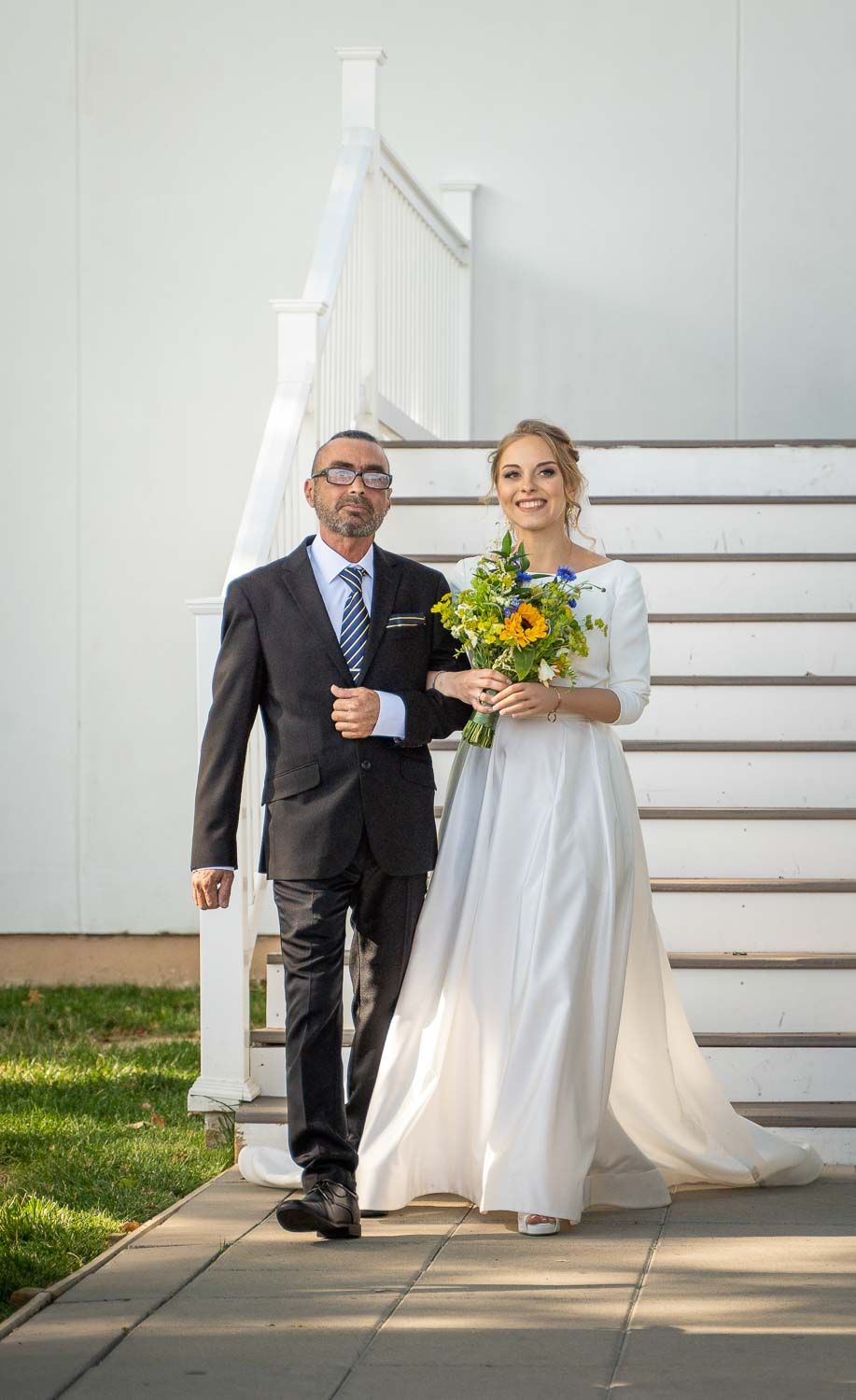 Bride in elegant white gown holding sunflower bouquet walks with father down outdoor wedding aisle.