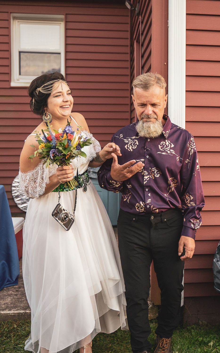 Smiling bride in white dress holding wildflower bouquet walking with bearded man in purple shirt.