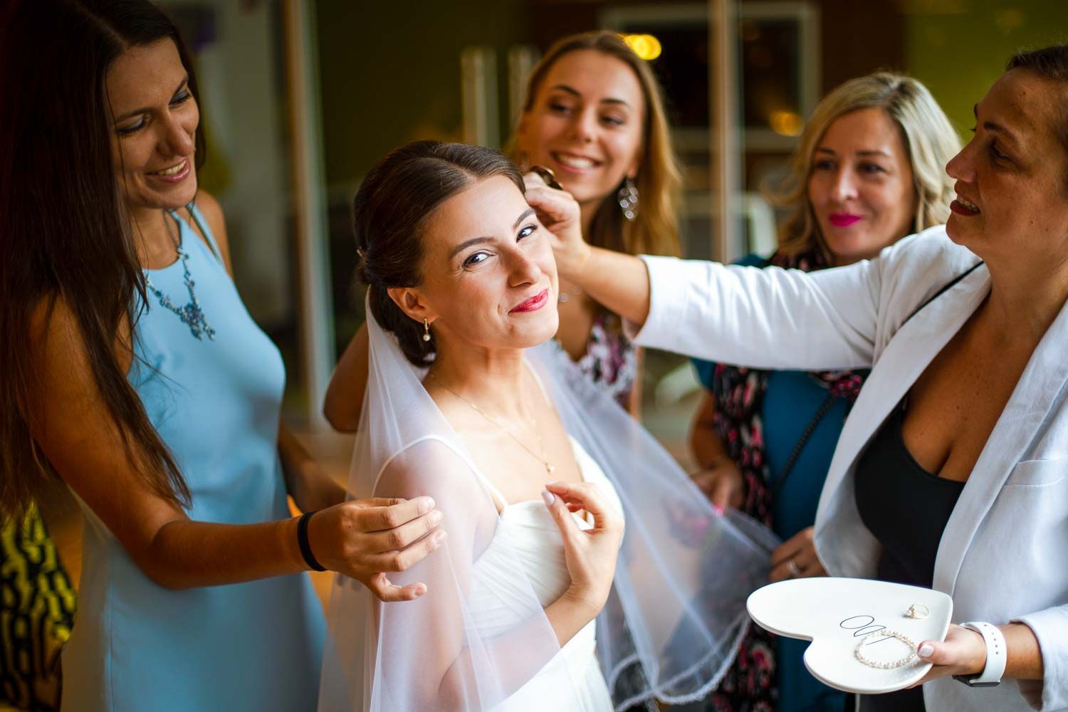 Bride smiling joyfully as bridesmaids in colorful dresses help with her makeup and wedding day preparations.