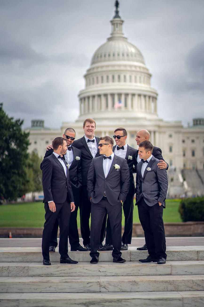 Groomsmen in black tuxedos gather around groom on US Capitol Building steps.