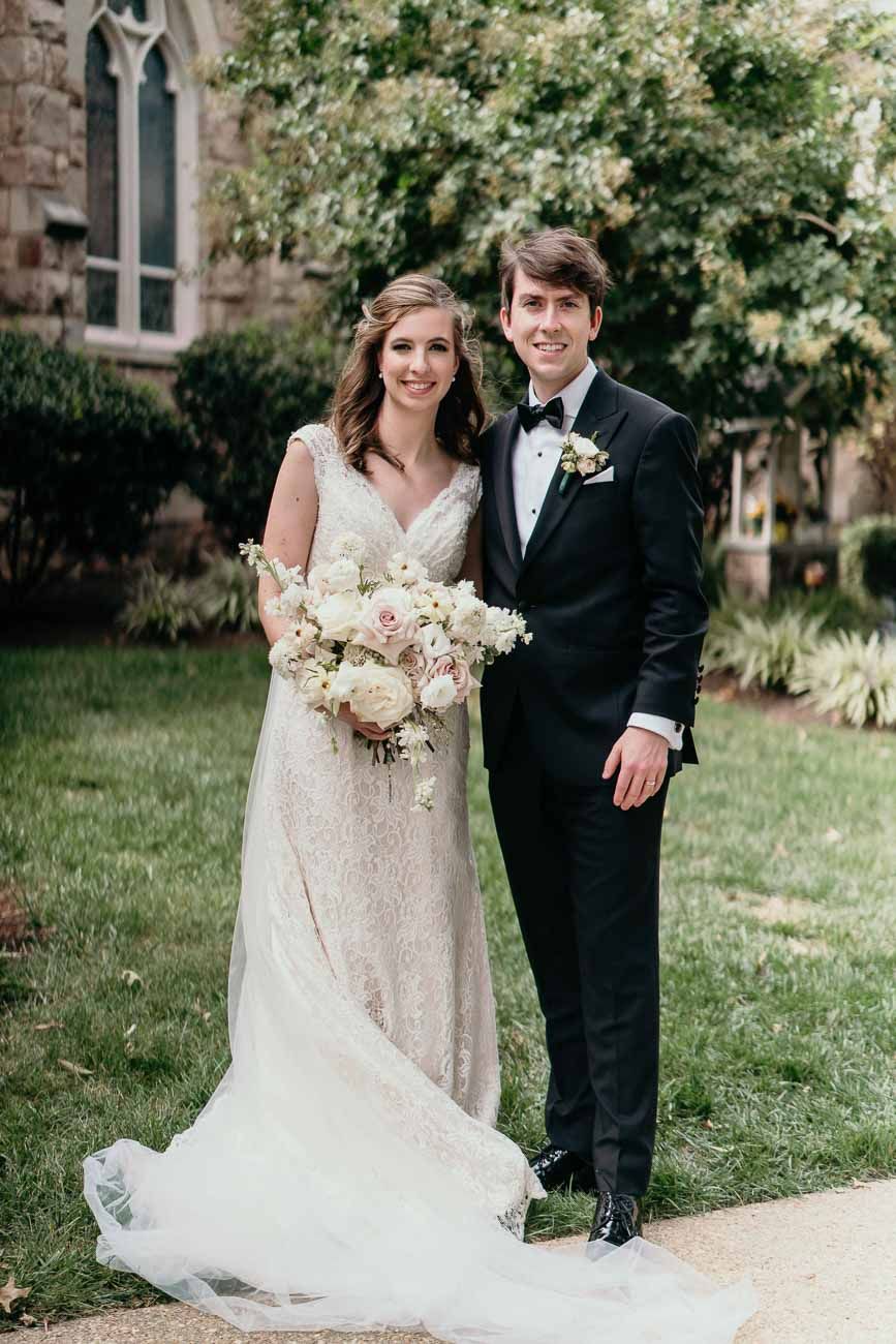 Bride in lace gown and groom in tuxedo stand together outdoors holding a romantic white floral bouquet.