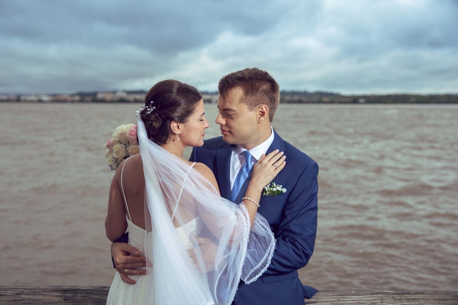 Romantic bride and groom embracing near water, bride in white gown with veil on cloudy day.