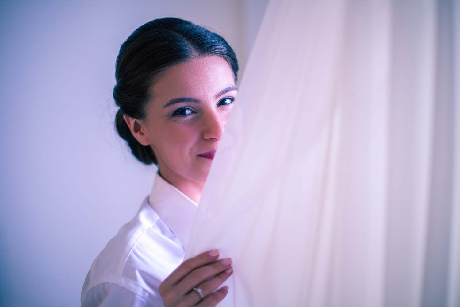 Elegant bride with an updo hairstyle peeking through a sheer white curtain before her wedding.