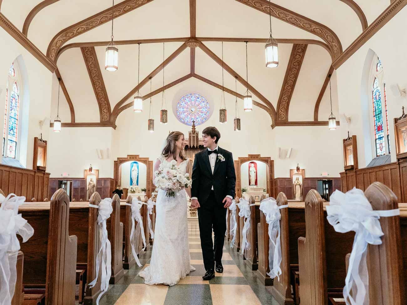 Bride and groom share a tender look in a sunlit church aisle with white bow-adorned wooden pews.