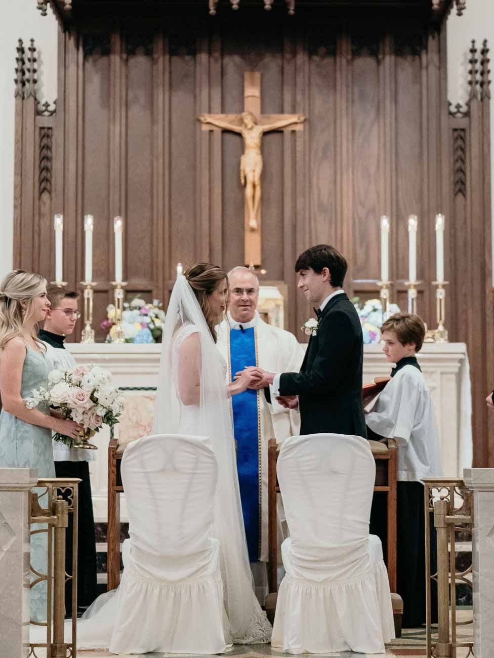 Bride and groom exchange rings at the altar surrounded by wedding party in Catholic church.
