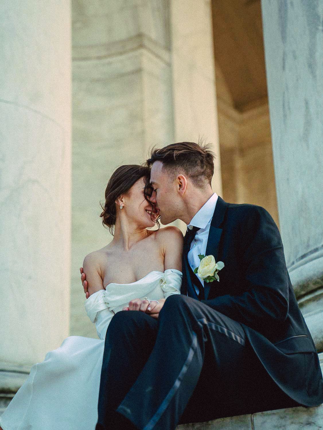 Bride and groom share an intimate moment seated between tall marble columns in soft golden light.