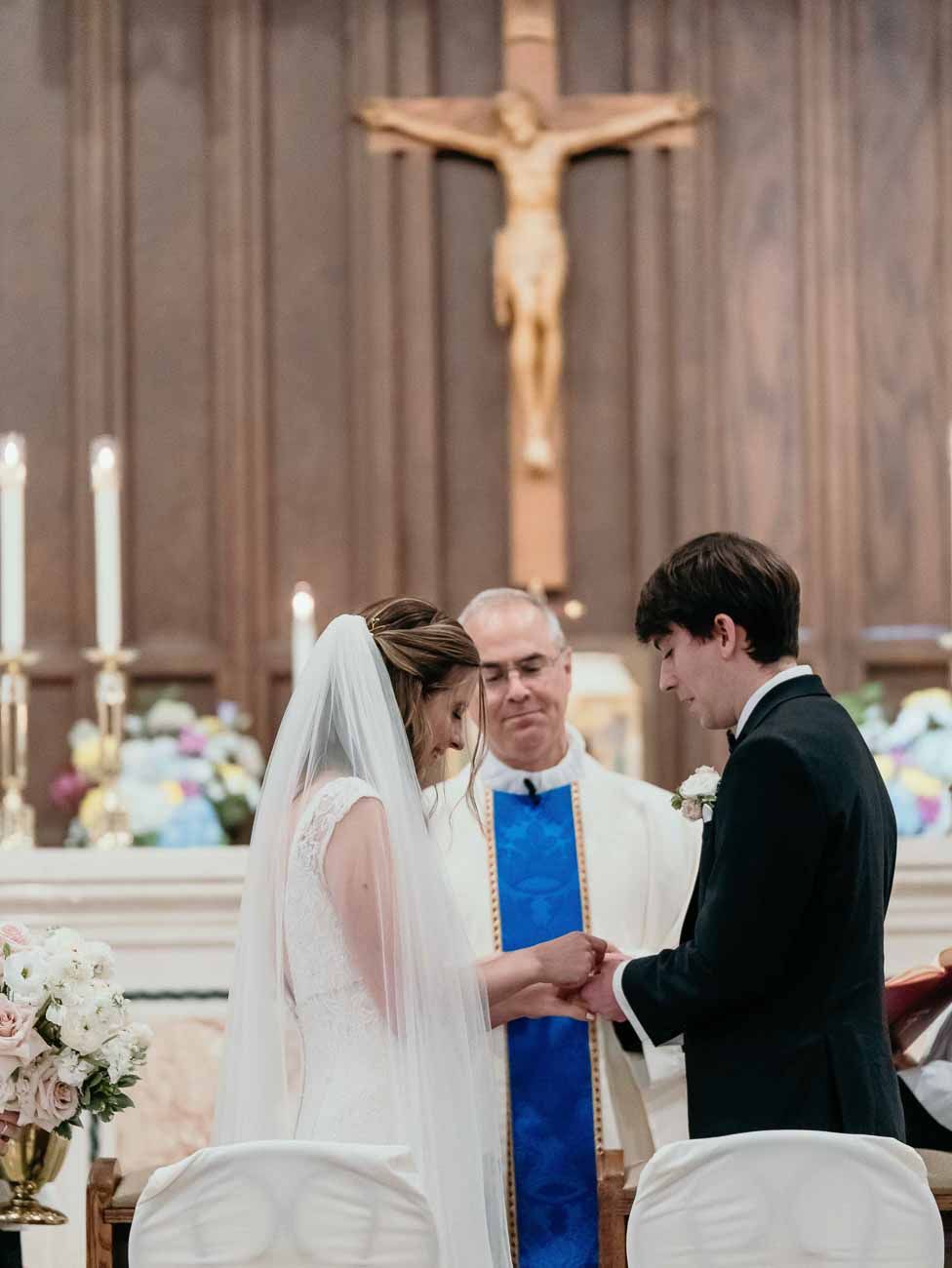 Groom places wedding ring on brides finger as priest watches during church ceremony.