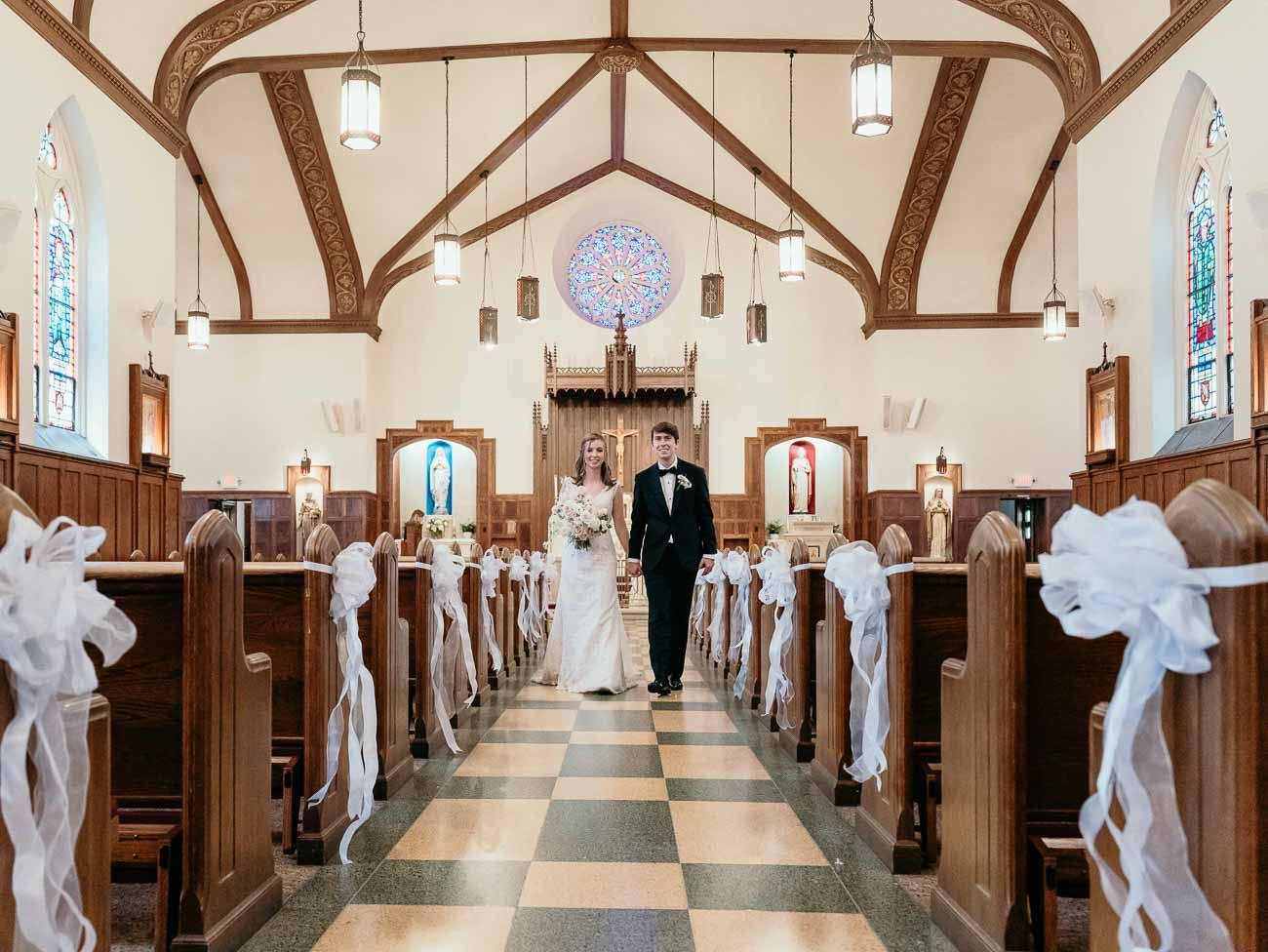 Newlyweds walk down a decorated church aisle with white ribbon bows and a rose window above.